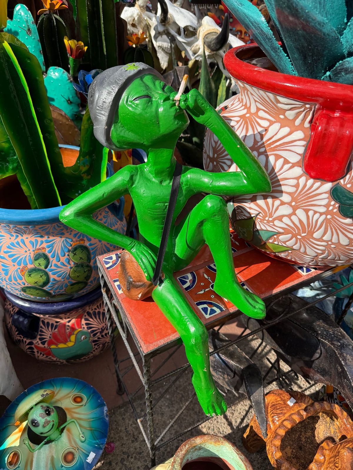 A garde statue of an alien wearing a beanie and smoking a joint sitting on a table. There are other brightly decorated garden pots and and metal cactuses around it. 