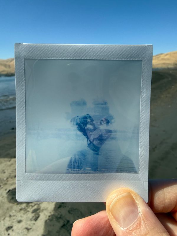 A hand holding an instant photo. There is dry ground and water in the background. The photo is faded, blurry layers of a figure, a tree, and a lake all together.