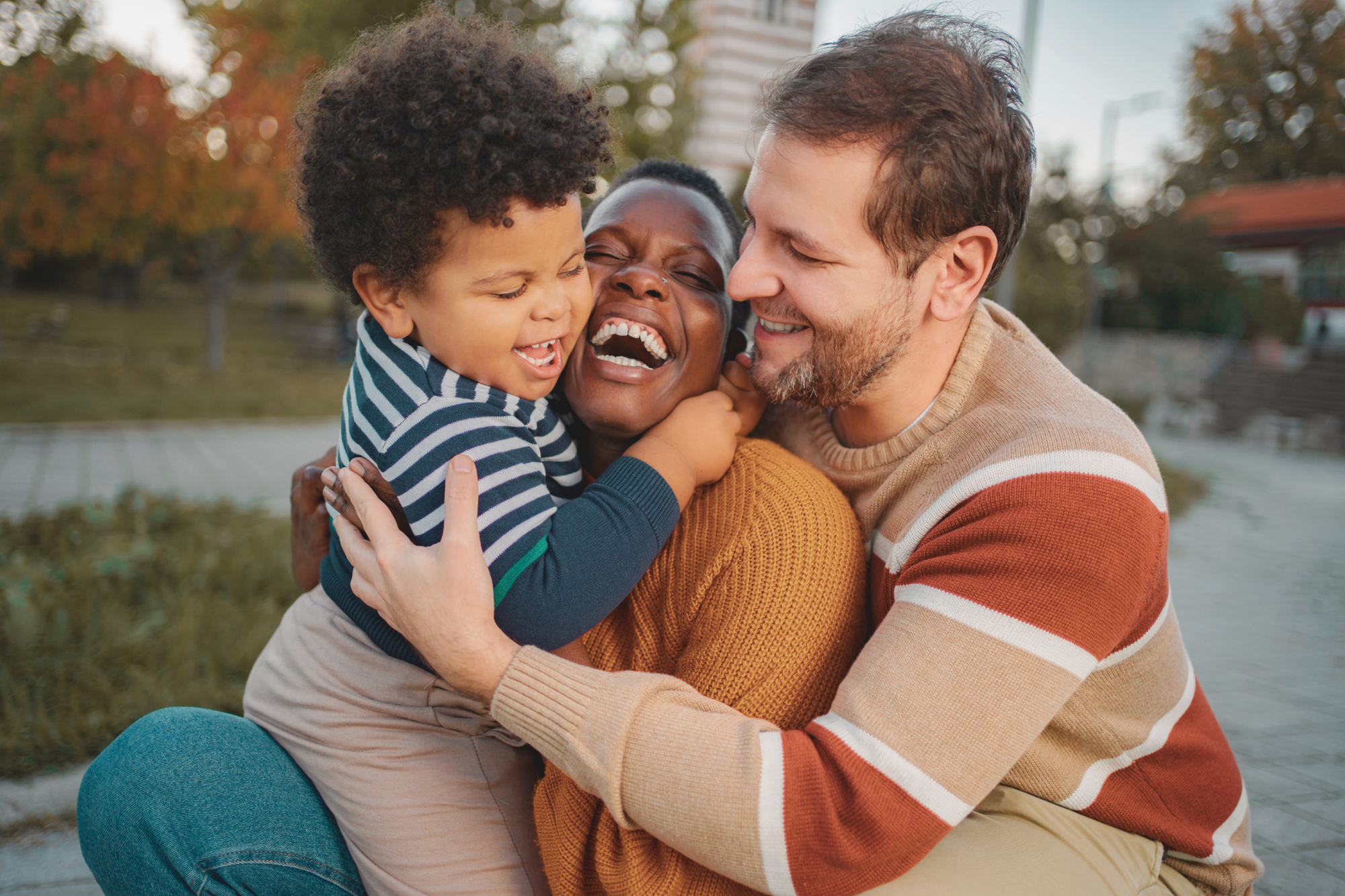 Toddler in stripped shirt hugging laughing Black mom in gold shirt while white dad in multicolored stripped sweater hugs the