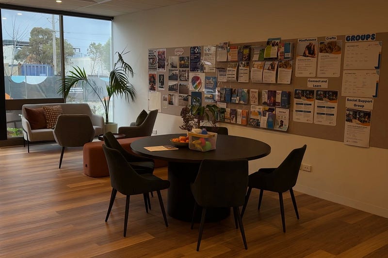 Photo inside the Brimbank Mental Health Connect Centre, where there are a table and chairs, a wall with educational pamphlets, and a couch and armchairs.