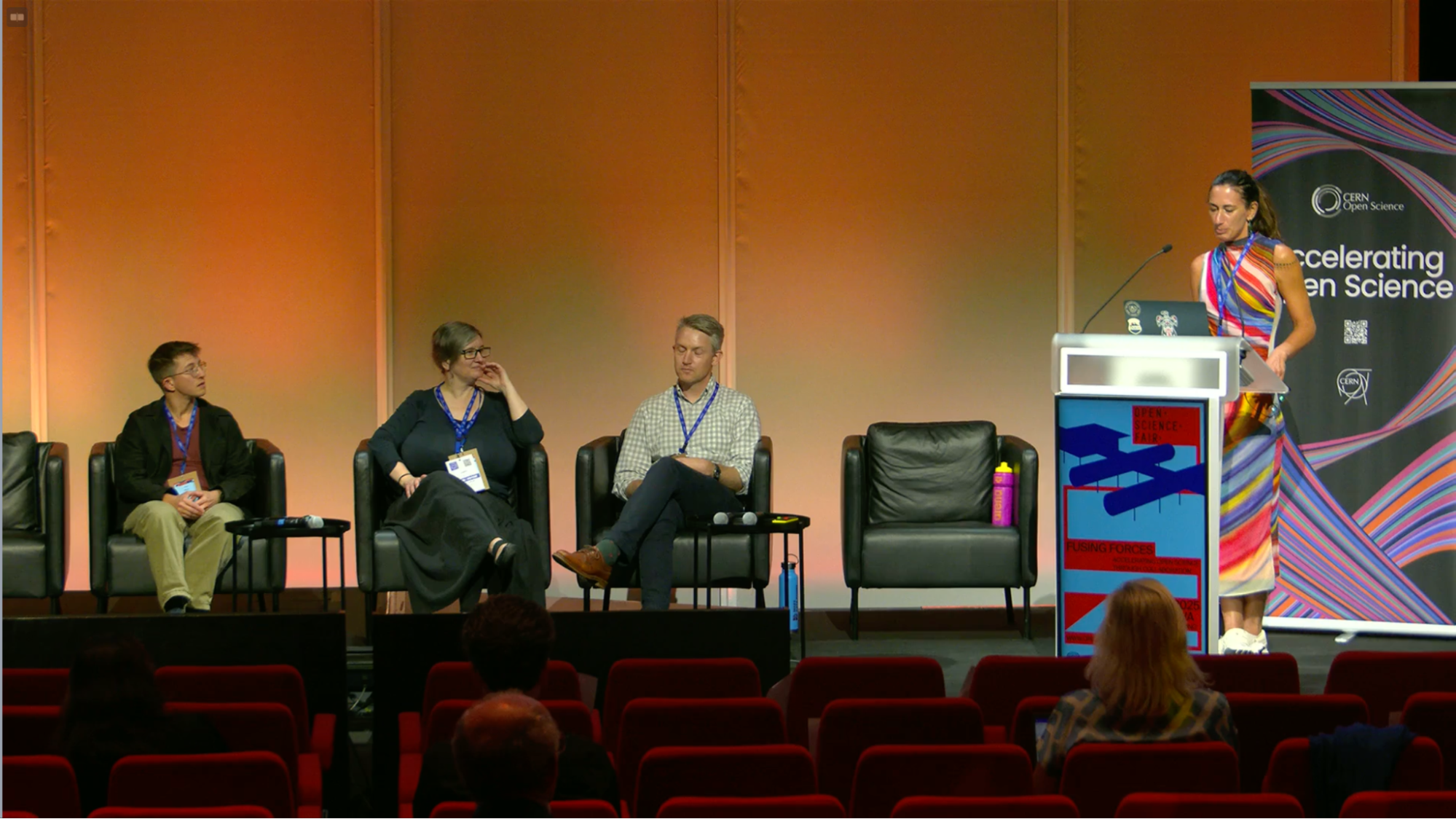 A woman in a colorful striped dress stands at a podium moderating a panel discussion at CERN's Open Science Fair. Three panelists sit in black leather chairs on stage - a man in casual attire on the left, a woman in dark clothing in the center, and a man in a light checkered shirt on the right. The backdrop features CERN Open Science branding with 'Accelerating Open Science' text and colorful geometric designs. Red auditorium seats are visible in the foreground.