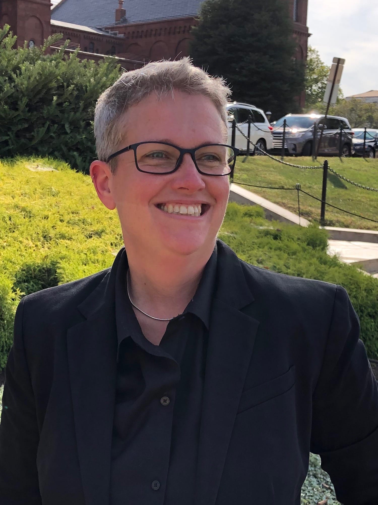 Photograph of Sharon Leon, a white person with gray hair and black square glasses, smiling, wearing a black button up shirt. In the background, a greenscape and a brick academic building.