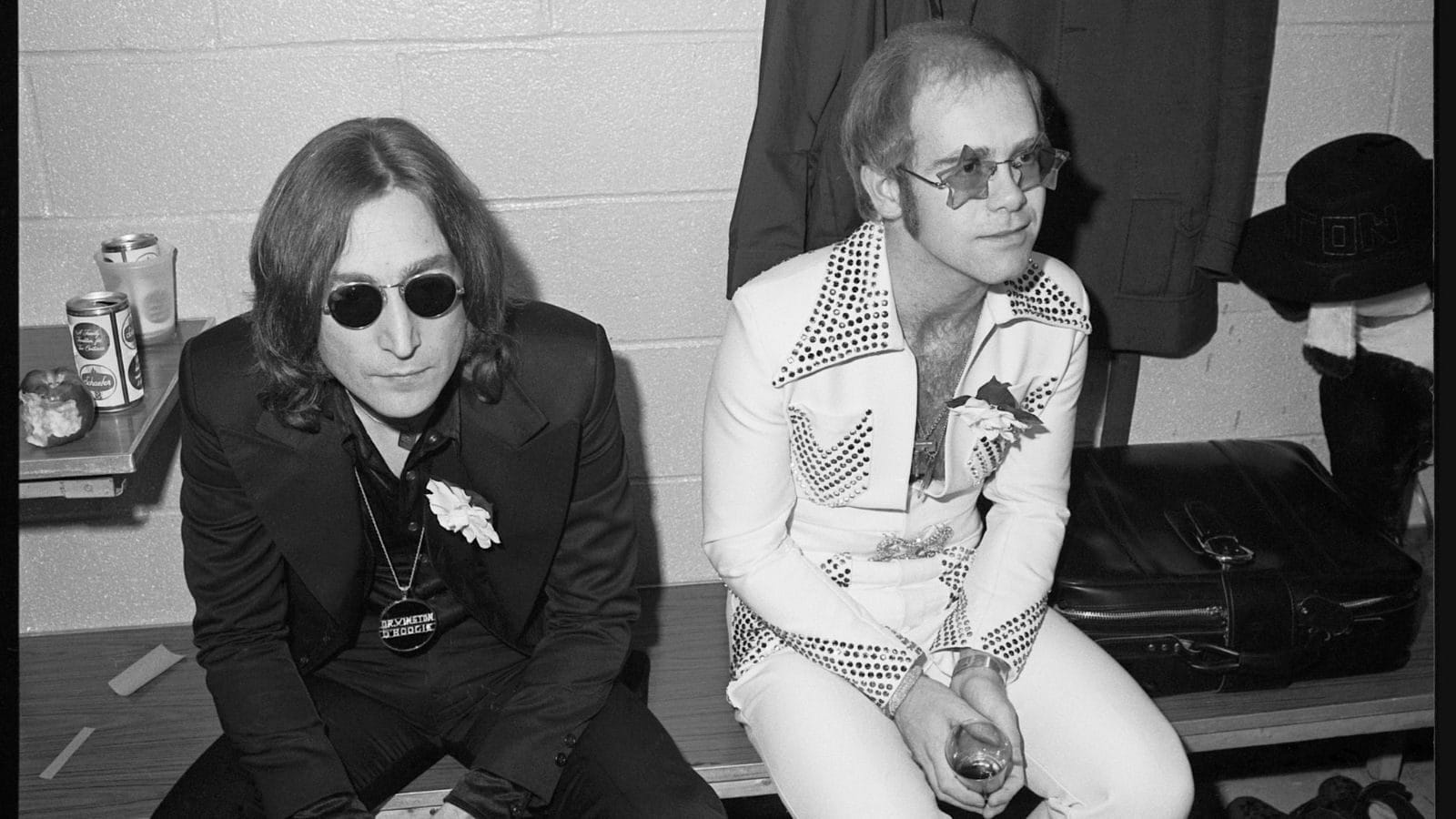 A black and white photo of John Lennon and Elton John, sitting backstage, looking very stiff and weird