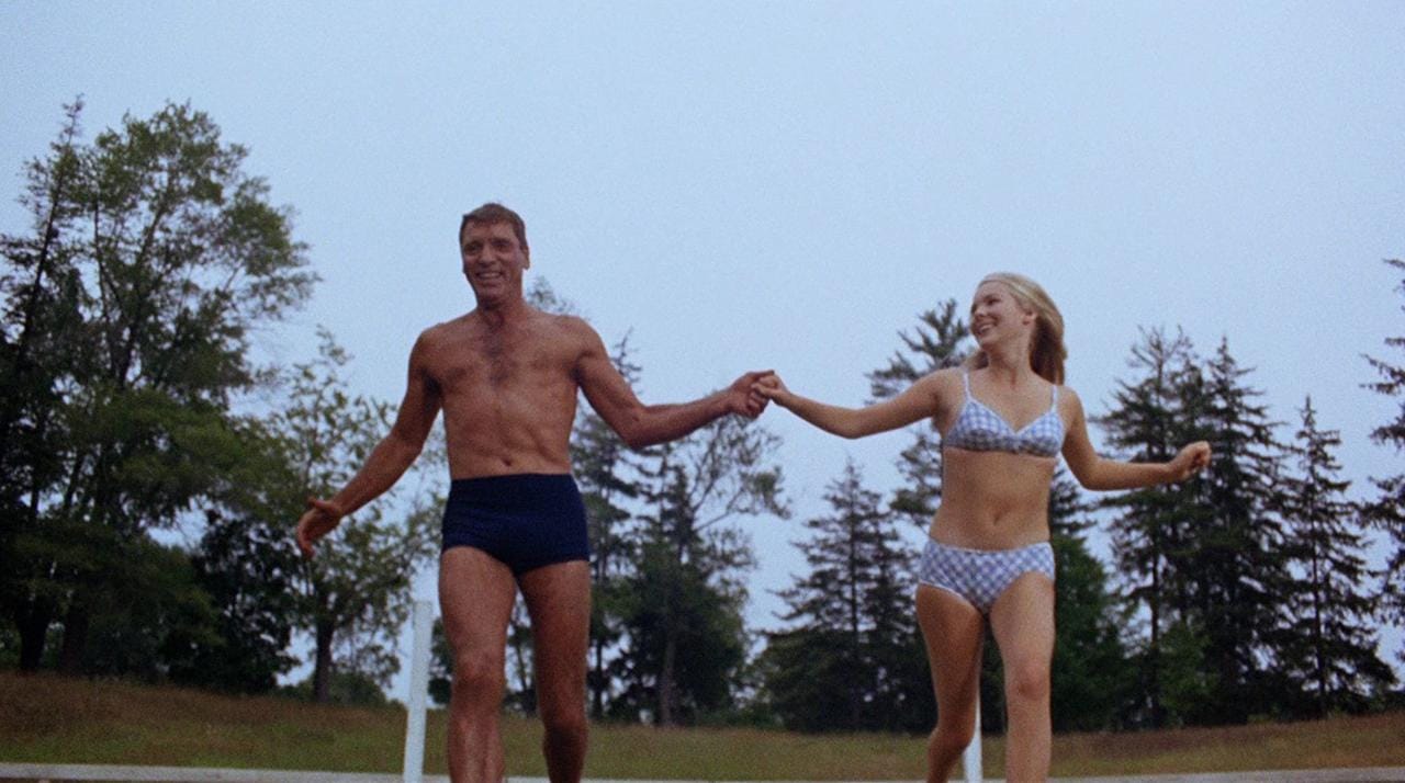 A still from The Swimmer showing an older Burt Lancaster in his little swimming trunks, frolicking in a field with his 21-year-old former babysitter, in a cute little bikini 