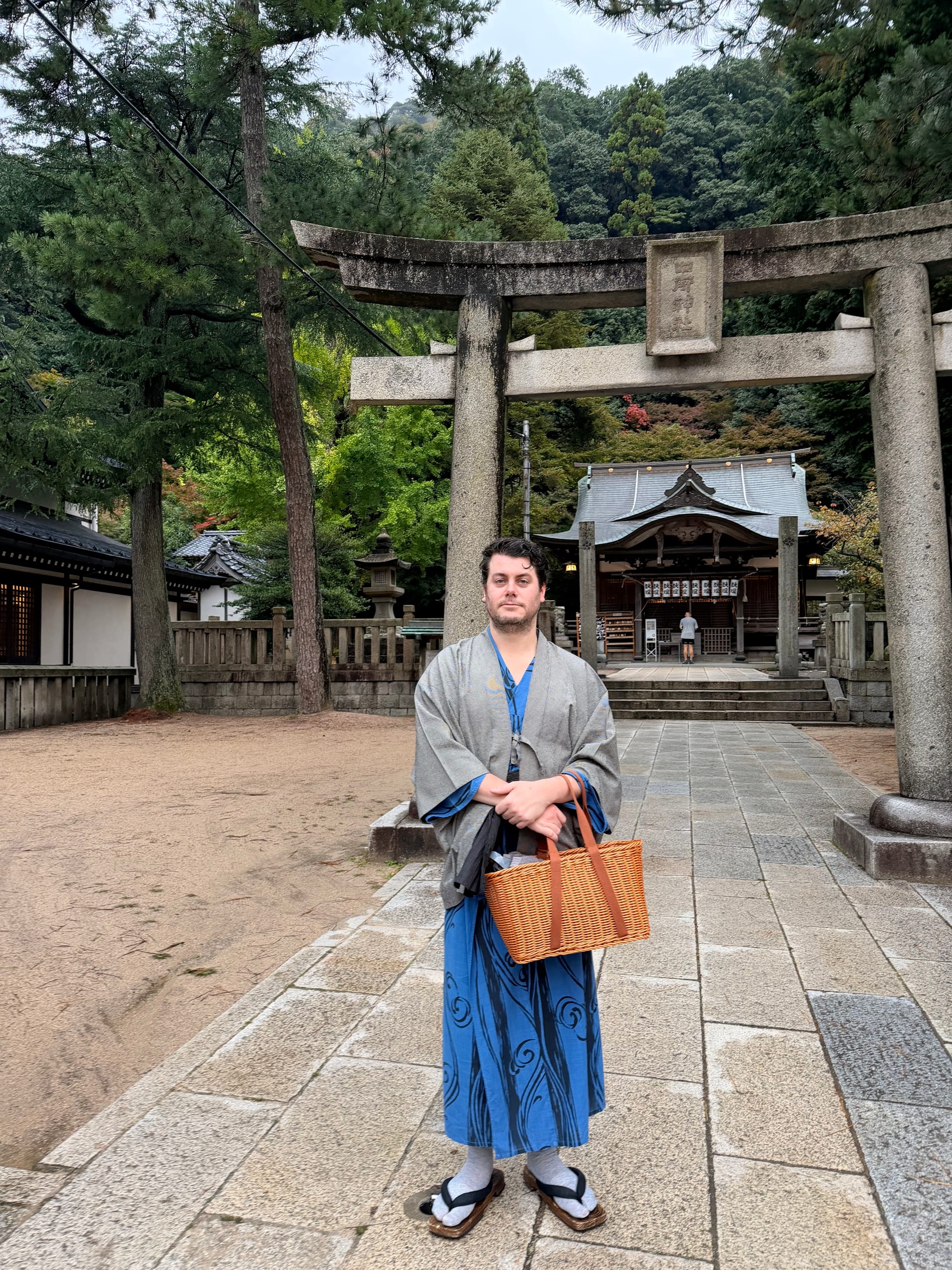 An image of me, wearing the traditional yukata — including the wooden sandals — standing in front of a temple. i'm also holding a wicker basket that they gave me, and I'm looking quite uncomfortable at being a white dude in this traditional Japanese clothing