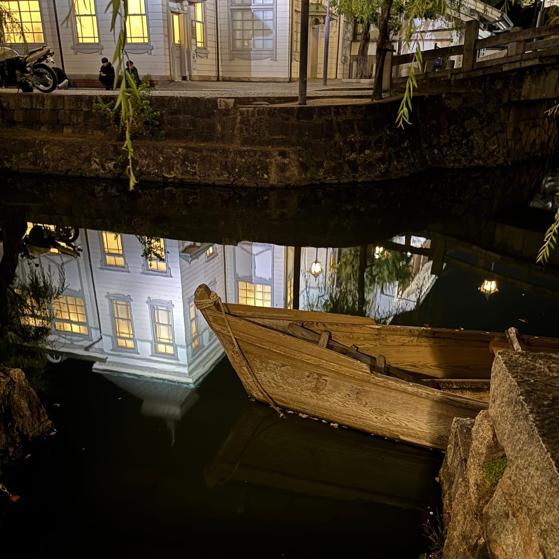A photo from the town of Kurashiki — a wooden canoe sits in the still water, which is reflecting an image of an old wooden building, lit up in the night