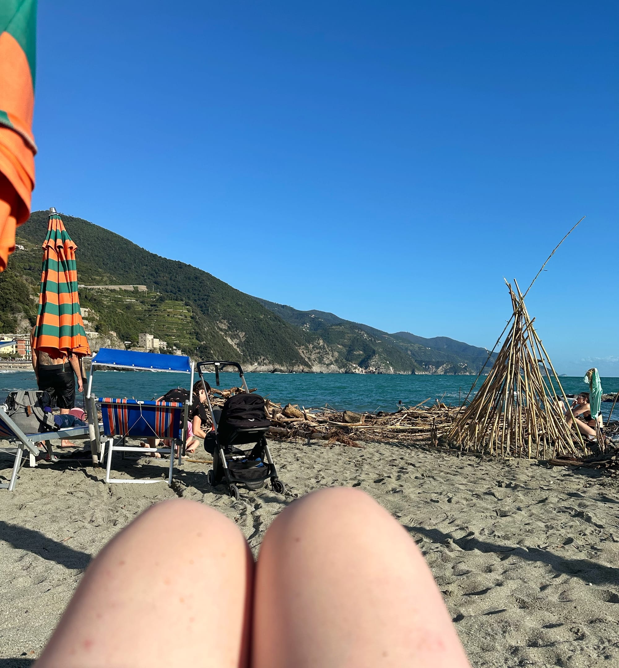 The author's knees set against the pristine blue sea of the beach in Monterosso. To the left, charming beach umbrellas. To the right, an impressive driftwood shelter built by random children. Idyllic hills in the background, clear blue skies above. 