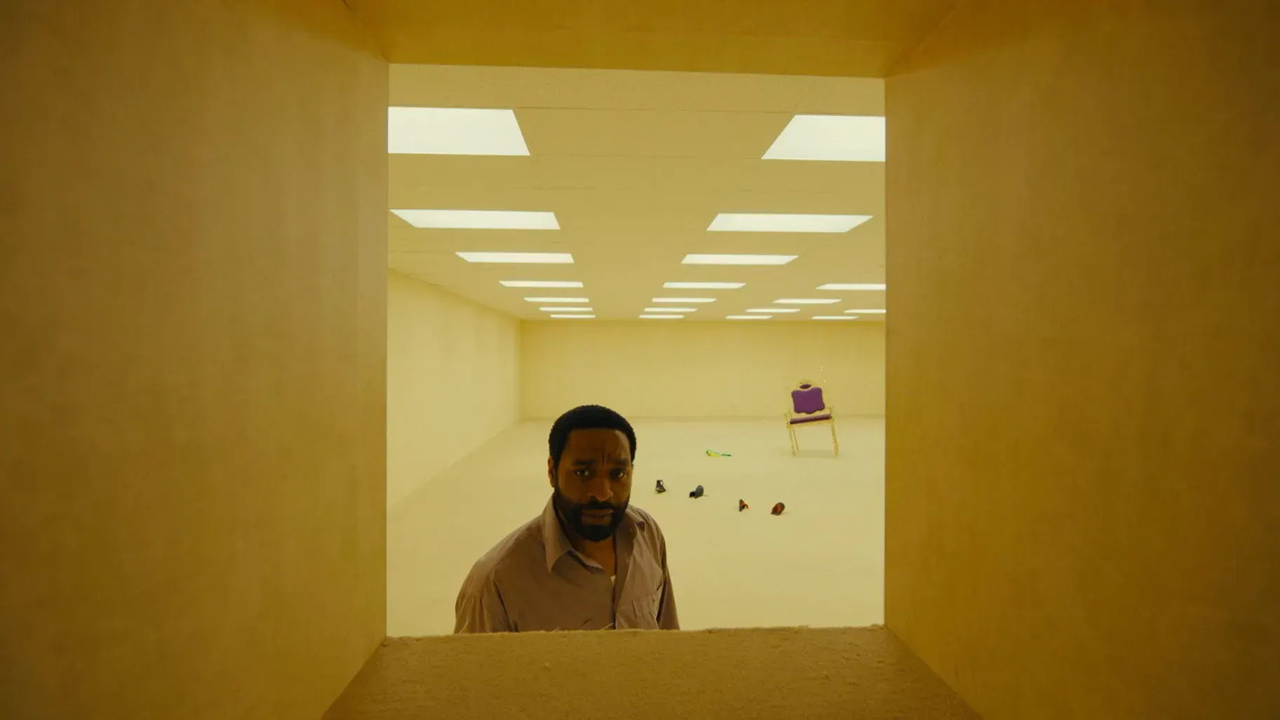 Chiwetel Ejiofor, a black man with a beard wearing a button up shirt, stands in a vast yellow room with fluorescent lighting looking into a square hole in the wall