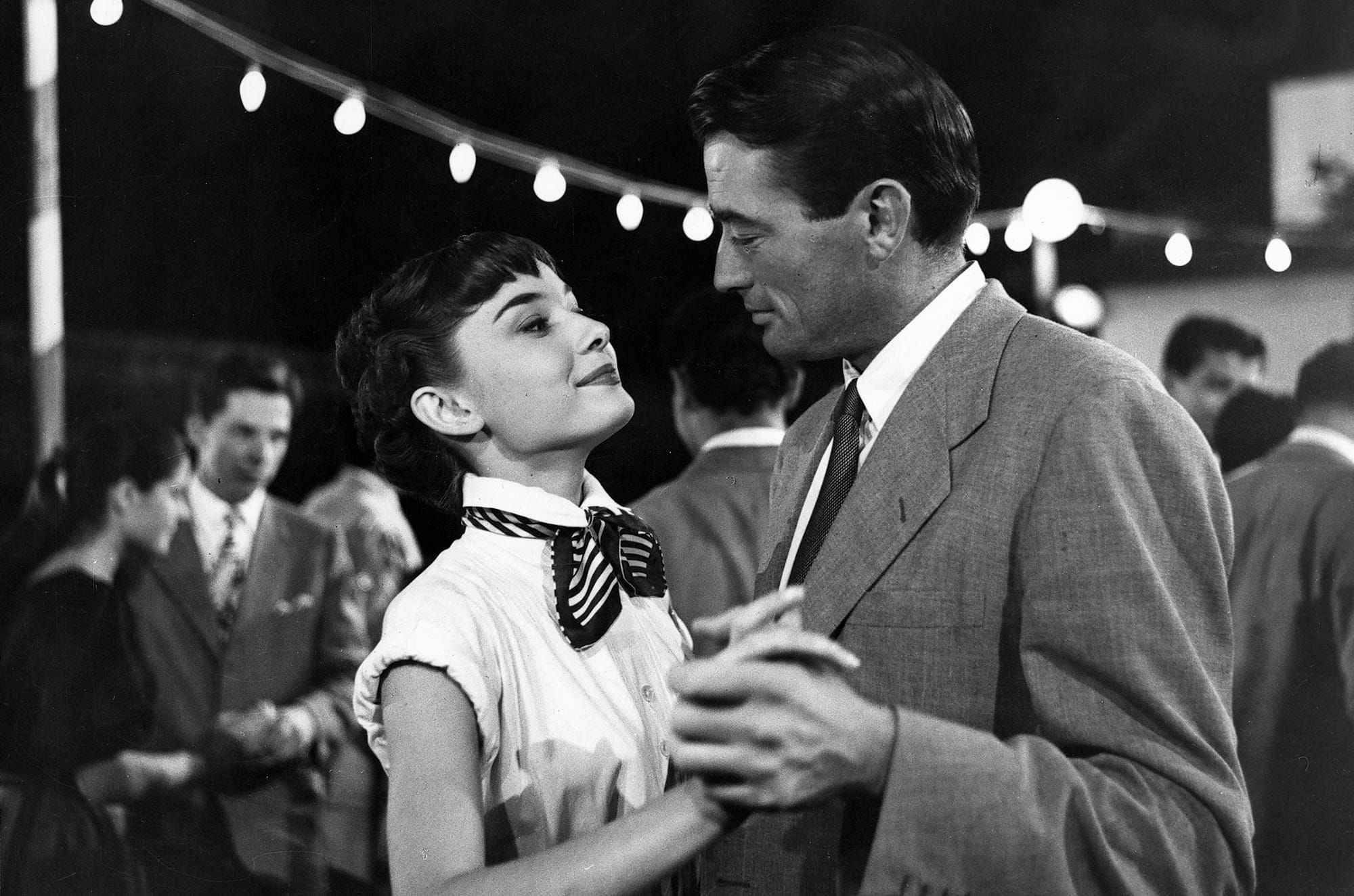 Audrey Hepburn and Gregory Peck dance outside in front of a string of lights