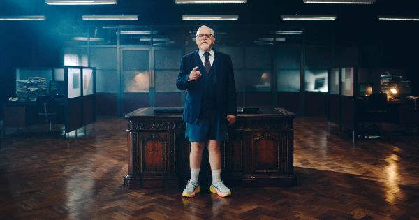 Brian Cox in his depressing ad for Asics, standing in front of a mahogany desk in shorts and sneakers