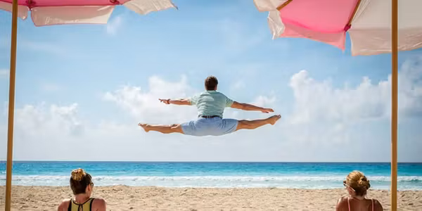 A man does a side split jump with his back to the camera over an idyllic beach while two women sit under beach umbrellas