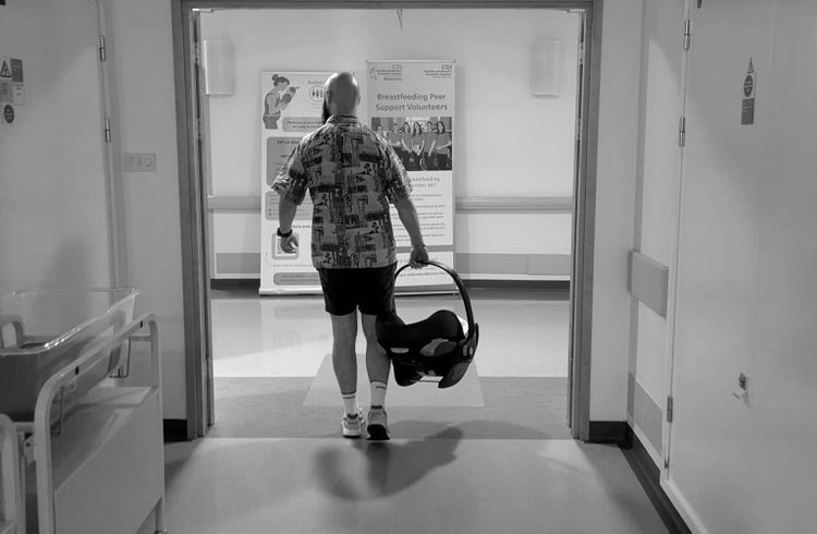 Bald man in a Hawaiian shirt walking along a hospital corridor with a baby in a carrier 