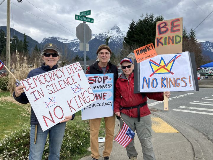 Three hold No Kings signs at the corner of Sauk and Seeman in Darrington, WA. Mountains in background. Democracy Dies in Silence, Stop the Madness, Be Kind.