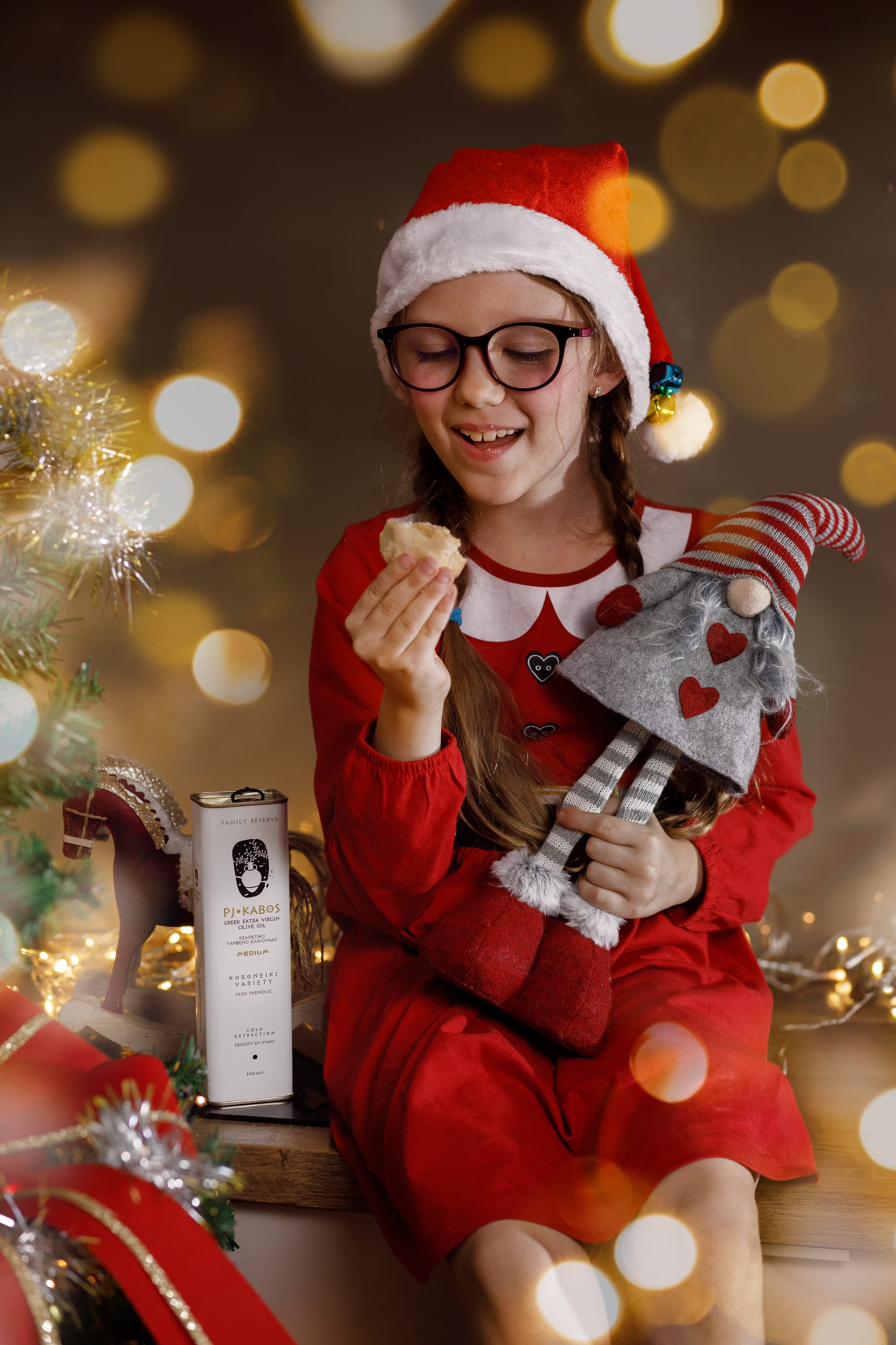 A picture of a little girl dressed as an elf at Christmastime holding a cookie she just baked with a tin of PJ Kabos Extra Virgin Olive Oil that she used by her side.