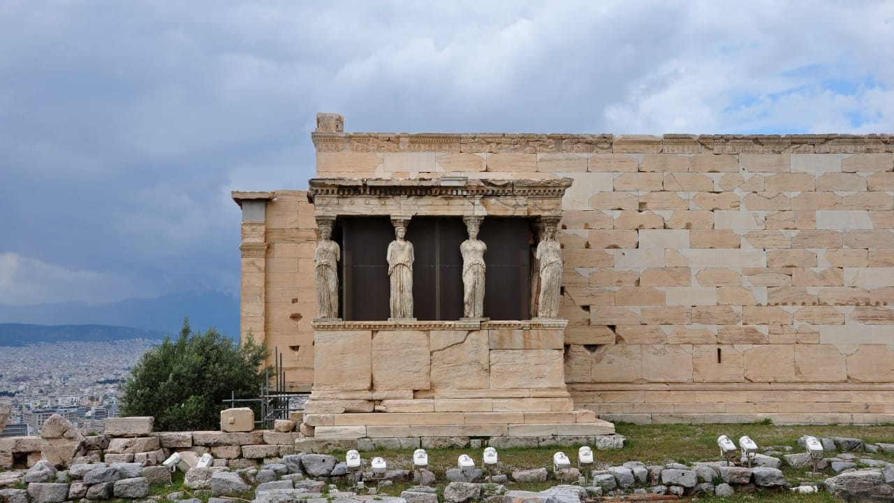 The Erechtheion's Porch of the Maidens (Caryatids), the top of the Sacred Olive Tree, and the sprawling city of Athens in the background.