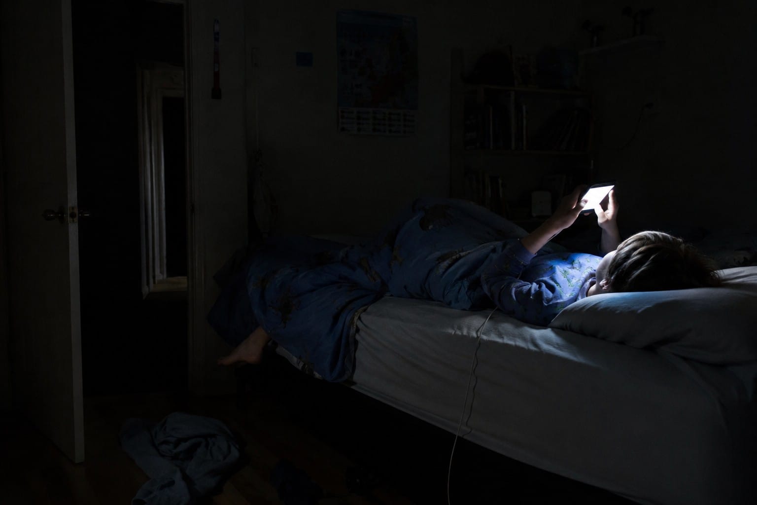 A child lying on a bed in a dark room lit only by a phone screen