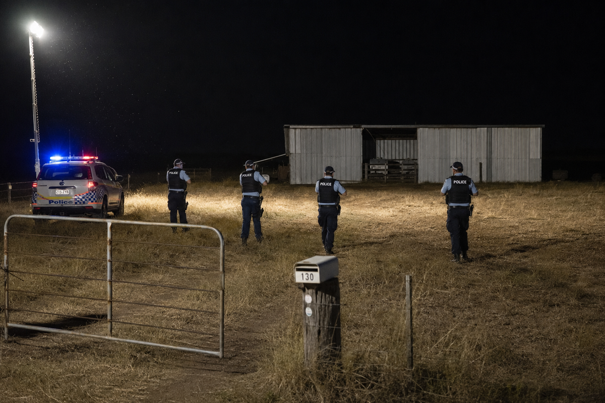 Queensland Police officers advancing across a floodlit rural paddock towards a shed at night