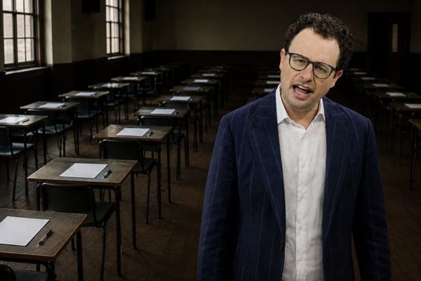 A man in a navy jacket standing in an empty examination hall lined with desks