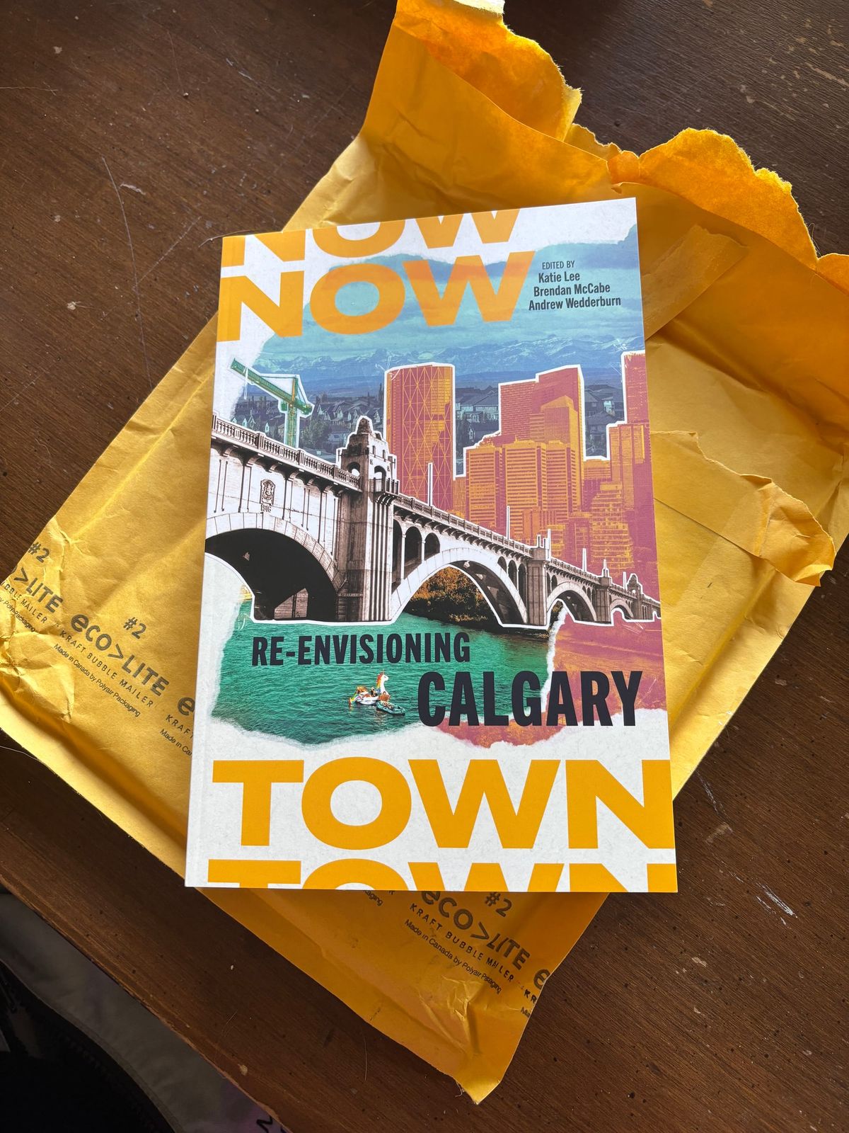 The book Nowtown lies on top of a mailing envelope on a wood table. The book has a colourful collage cover. The full title is Nowtown: Re-Envisioning Calgary.