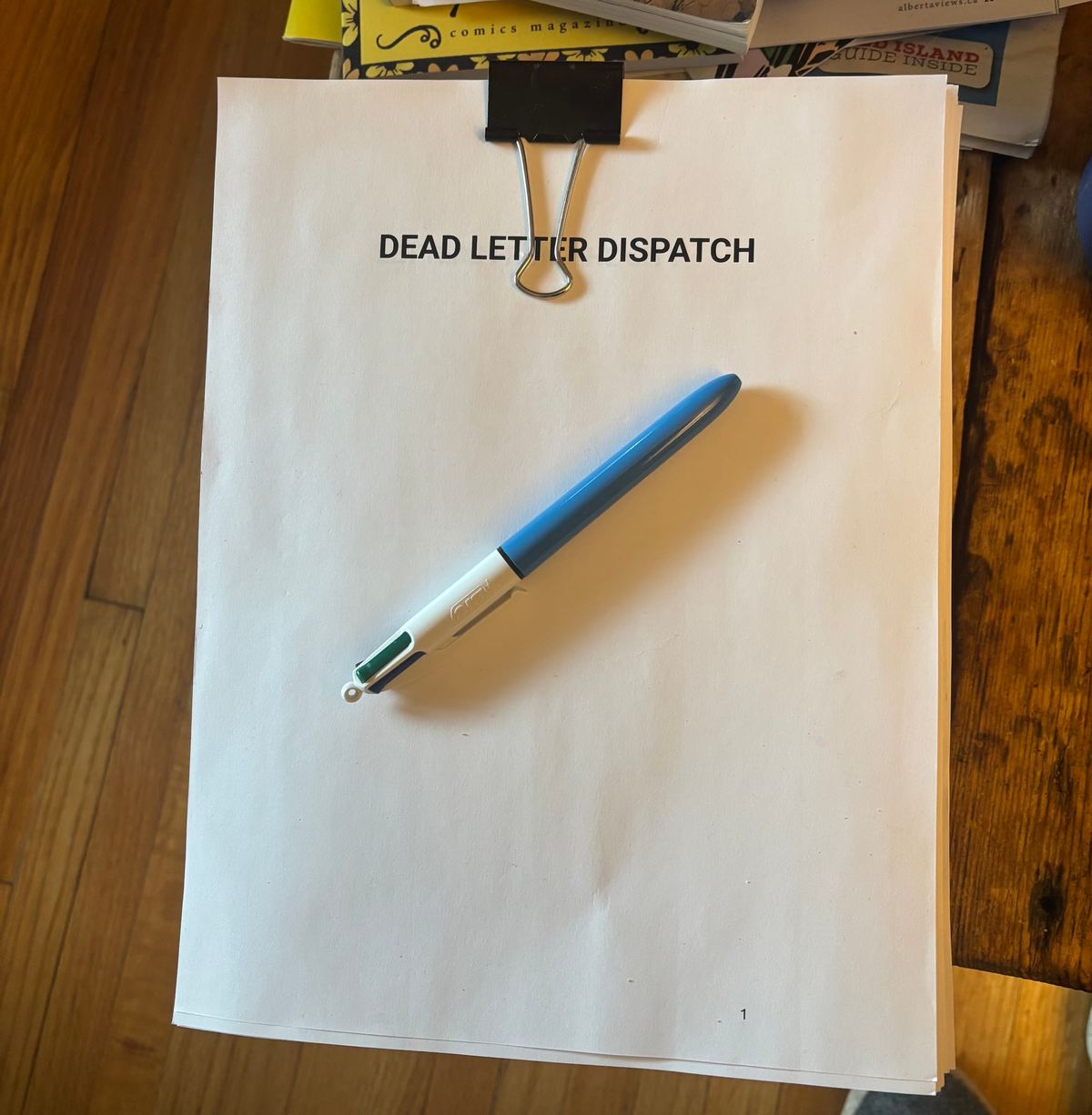 Printed pages from a manuscript on a wood table. The pages are held together by a a binder clip. The title of the manuscript is Dead Letter Dispatch. There is a Bic pen resting on top.