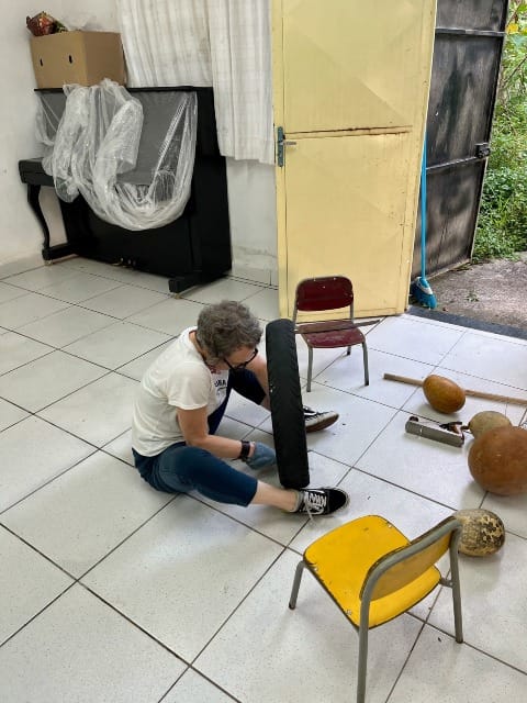 A woman sits on the floor holding an upright motorcycle tire between her feet. With her gloved hands she is using a big knife to cut away the rubber inside the tire to reveal the wire beneath. We see a few dried gourds on the floor and an open doorway onto a backyard.