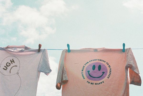 Two t-shirts drying. One saying UGH. Another with a happy smiley saying 'Don't want for everything to be perfect. Be happy'.