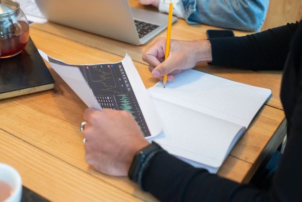 Close-up of hands writing notes on notebook during business meeting with laptop and charts.