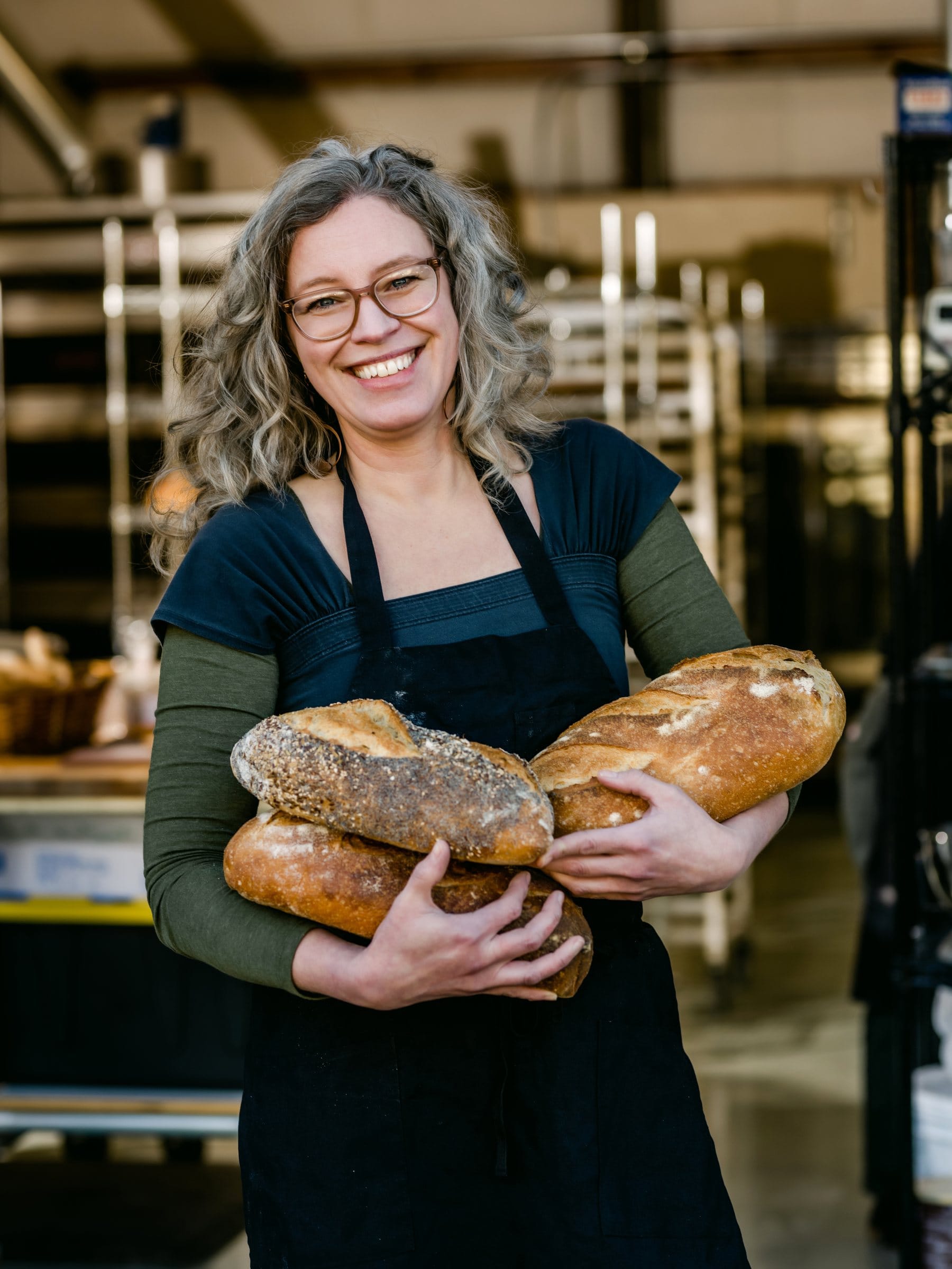 Baker in her bakery holding an armful of fresh sourdough loaves