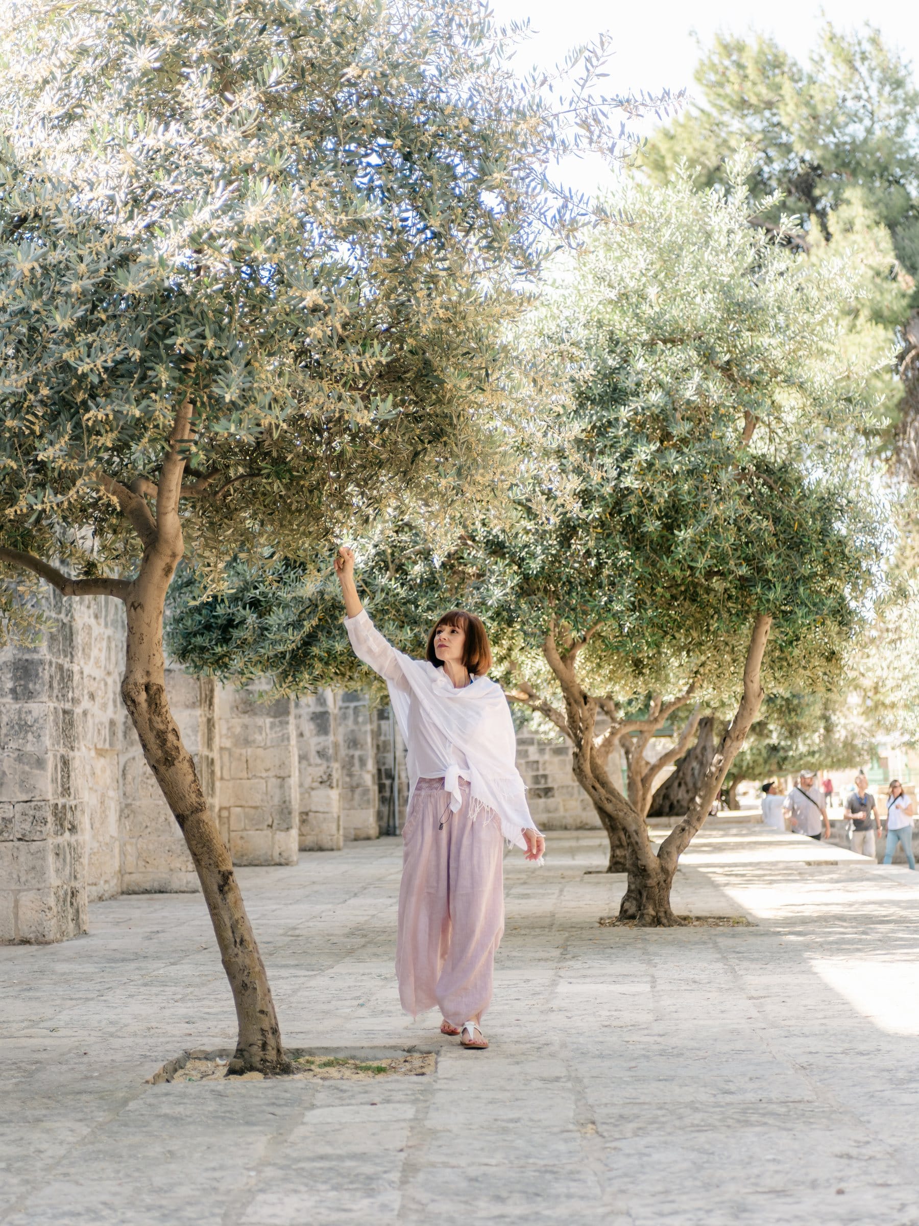 Woman twirling beneath olive trees beside an old stone wall