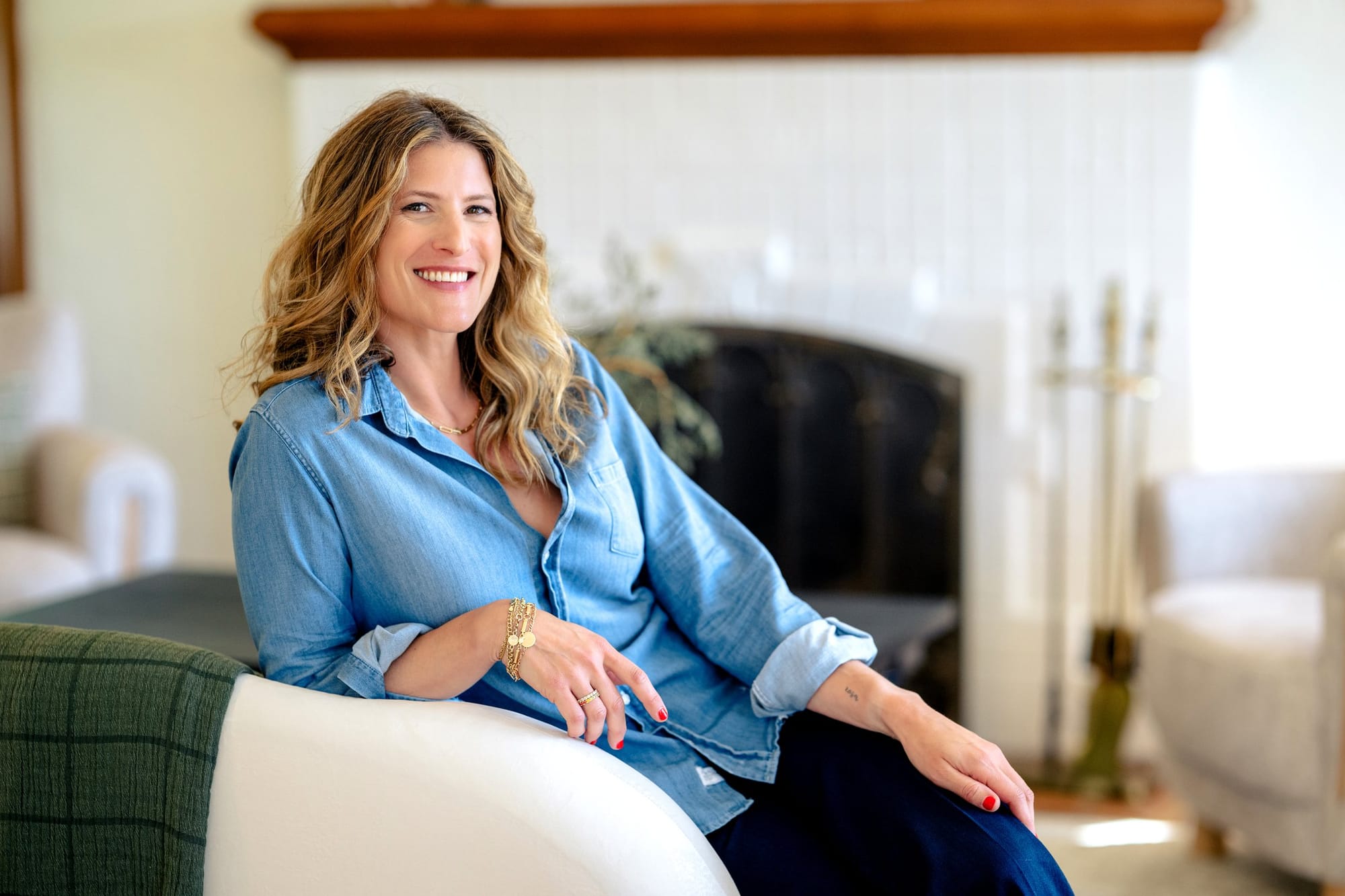 Woman in a chambray shirt smiling on a white sofa by the fireplace