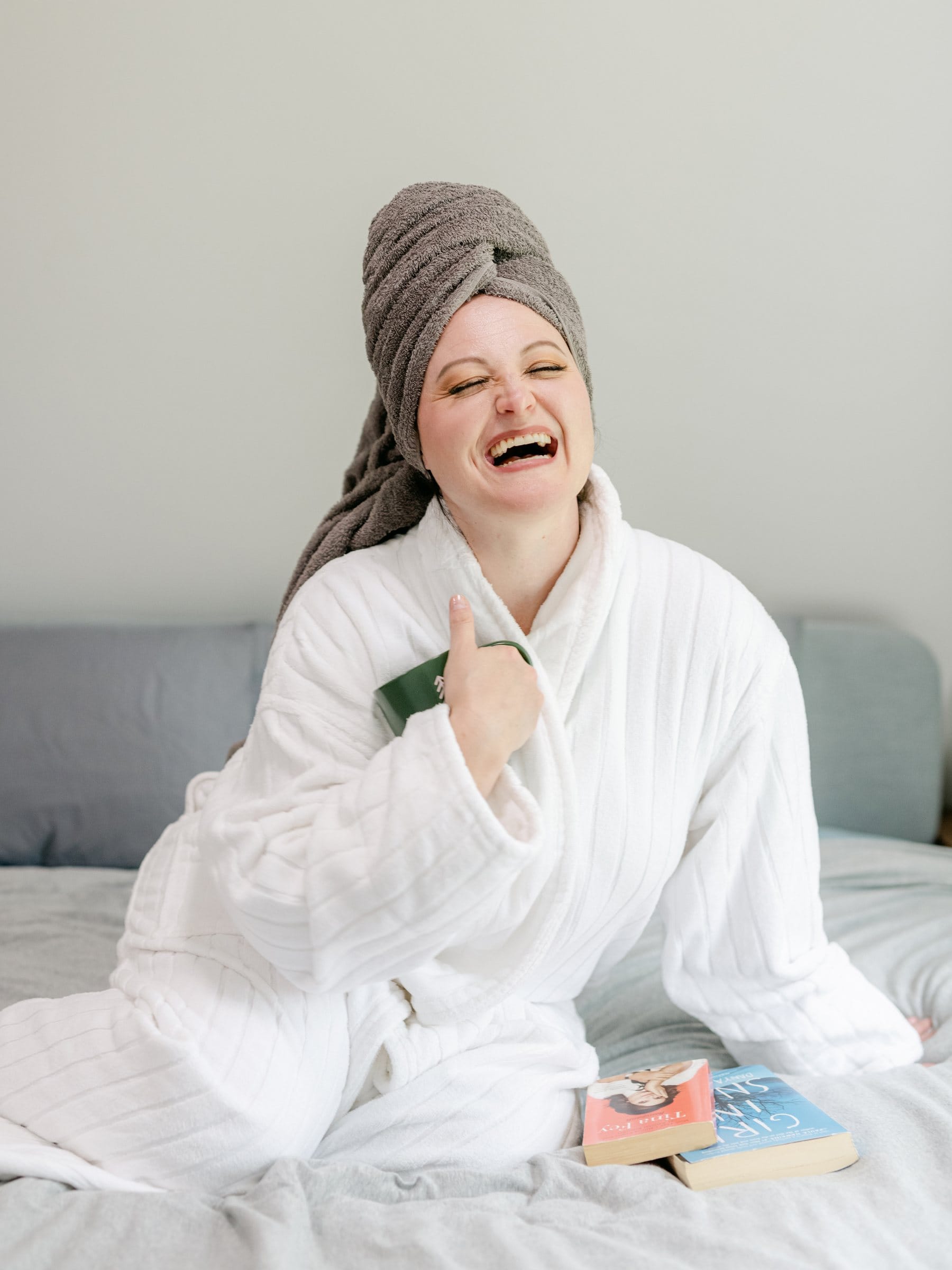 Woman in a bathrobe and hair towel laughing on her bed