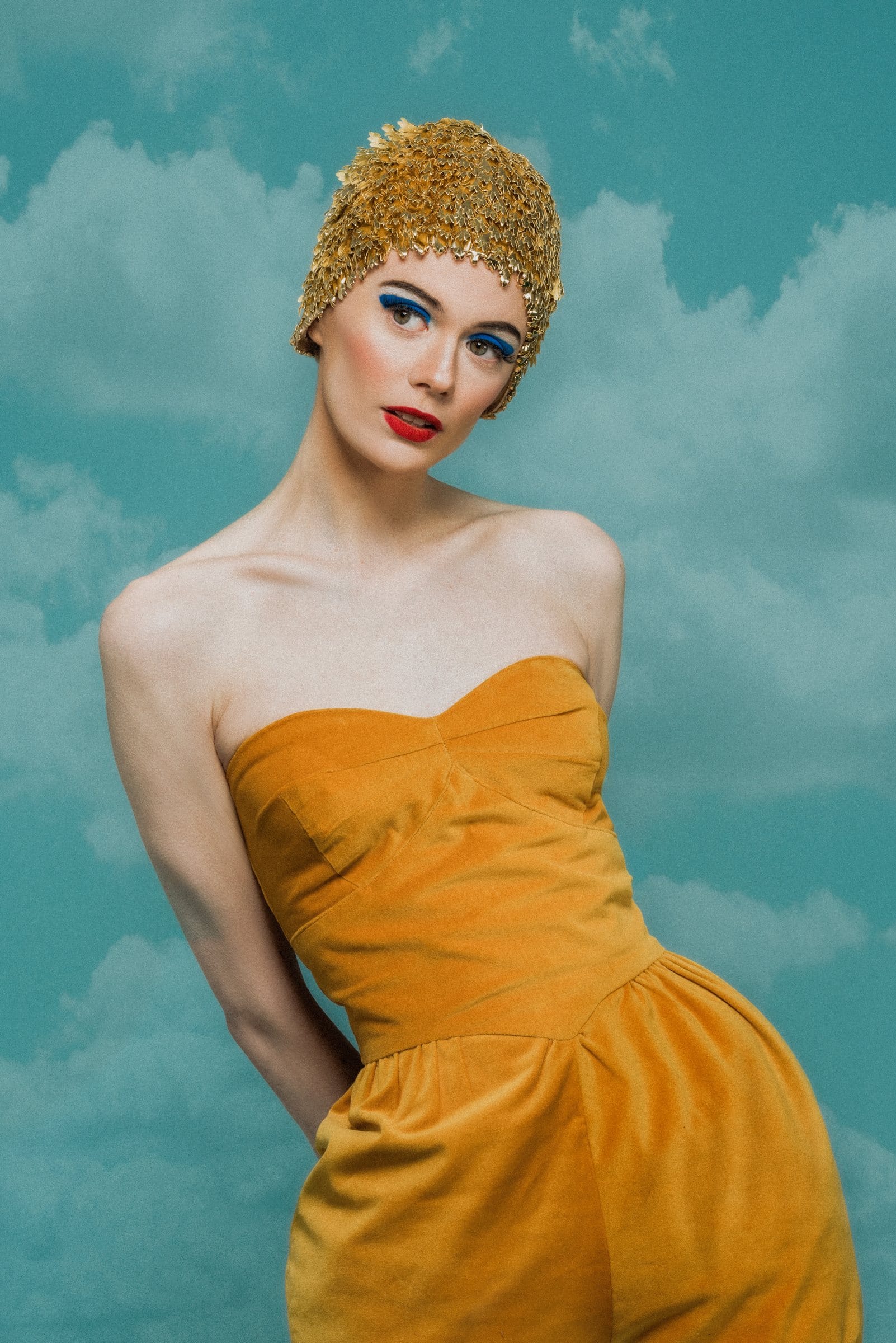 Editorial portrait in a gold cap and marigold dress against painted clouds