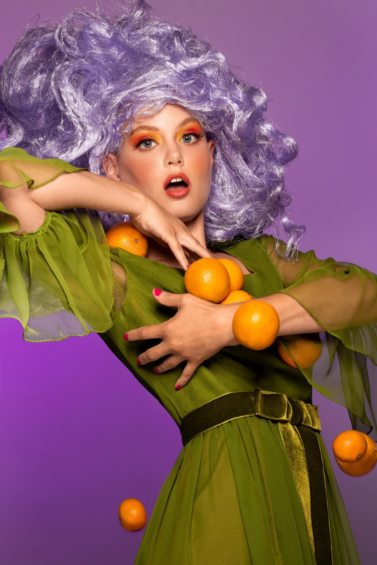 Editorial portrait with a lavender wig, green dress and tumbling oranges