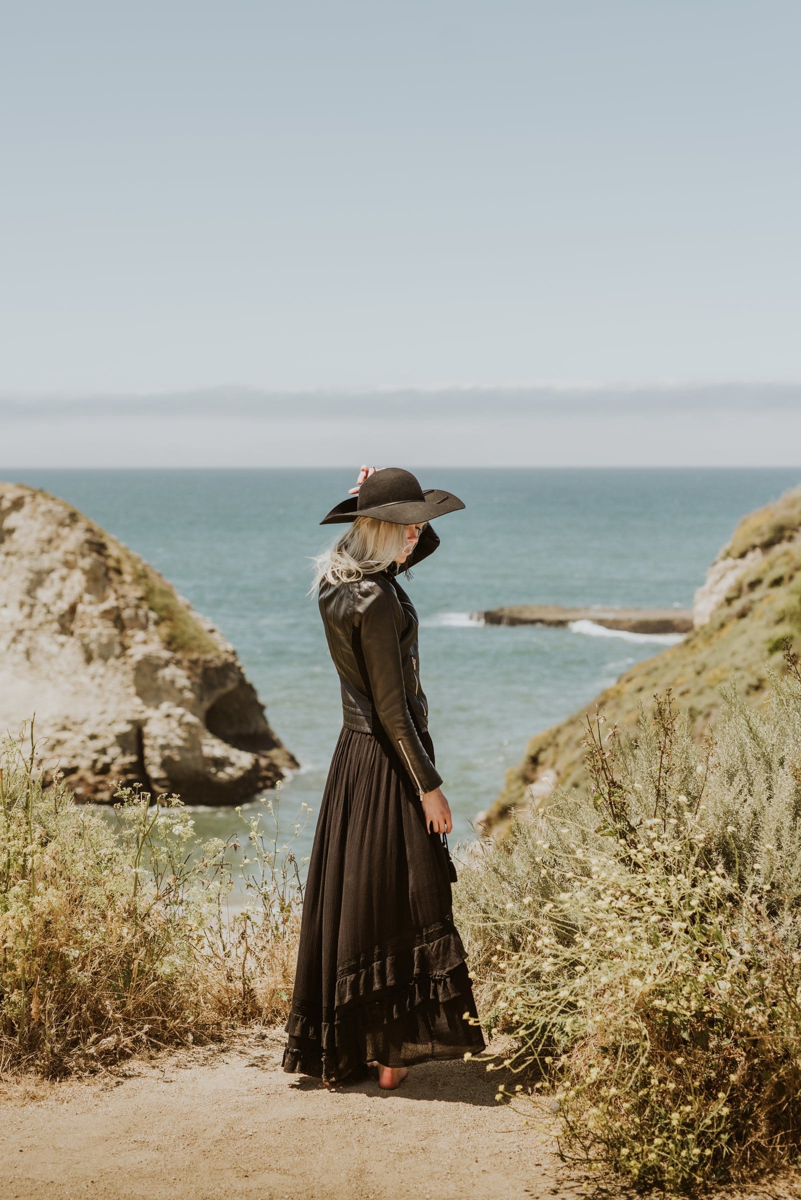 Woman in a black gown and hat looking out over a coastal cove