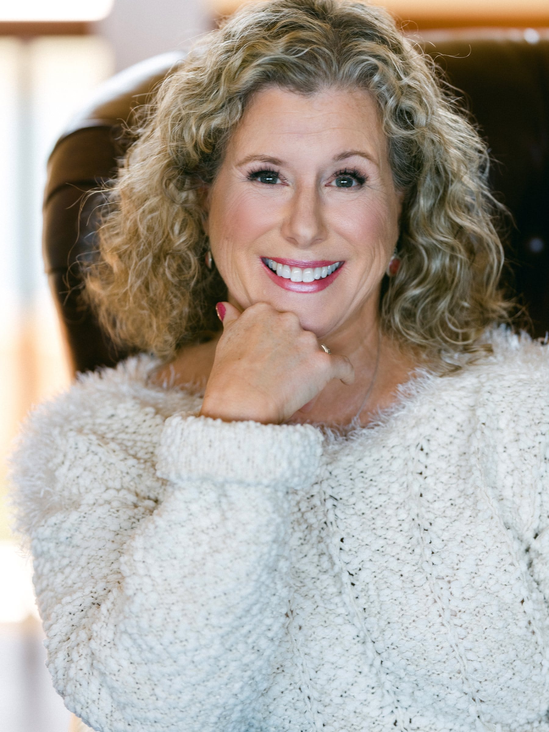 Curly-haired woman in a cozy cream sweater, chin on hand