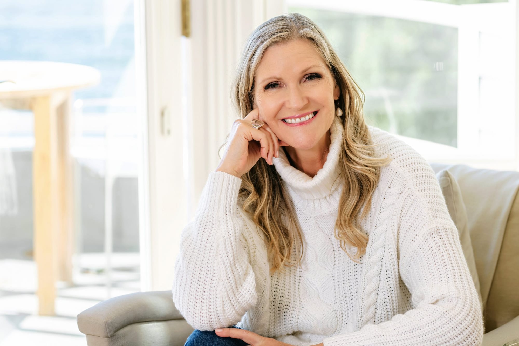 Woman in a cream sweater resting her chin on her hand in a sunny living room