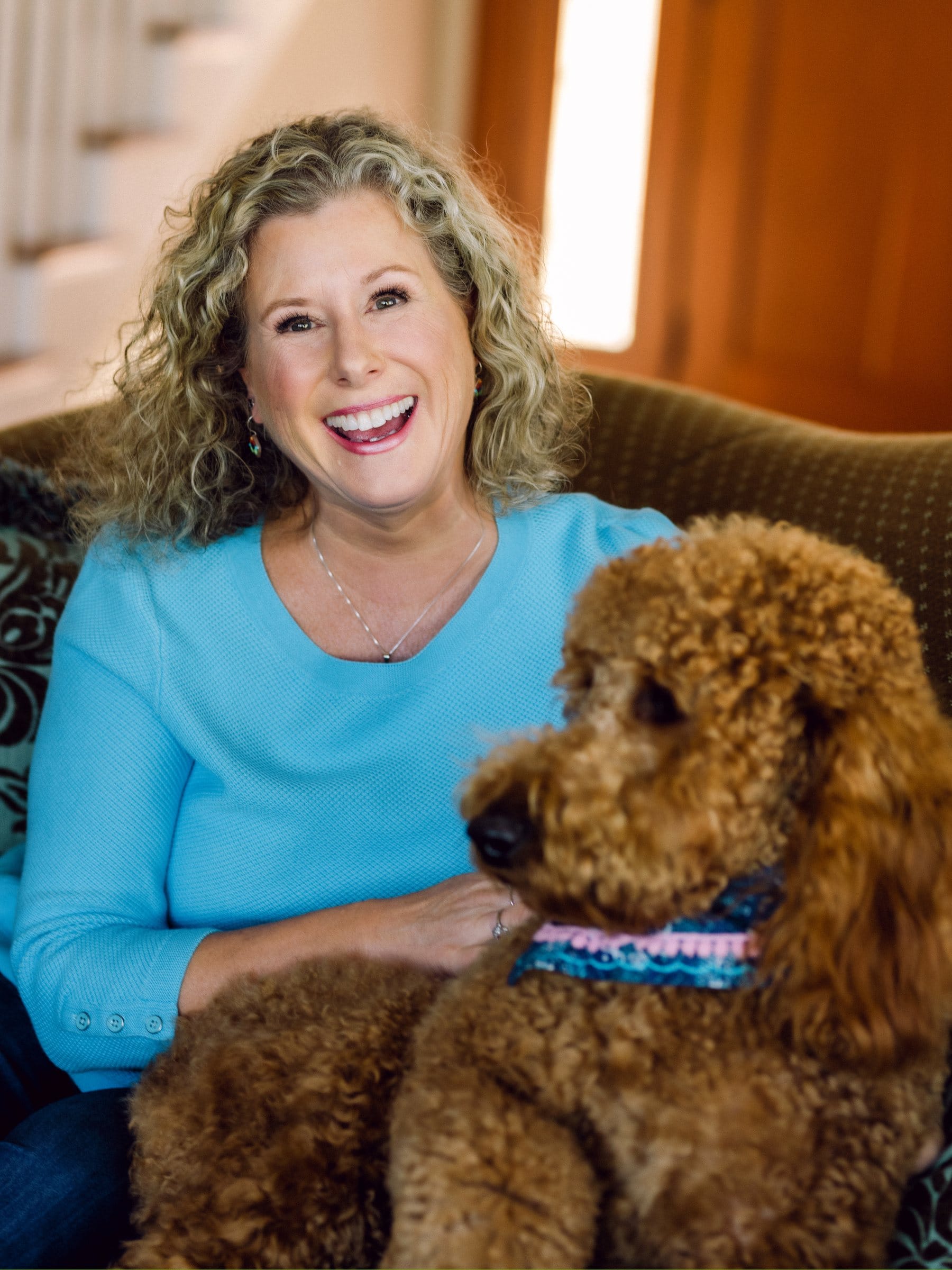 Curly-haired woman laughing on a sofa with her goldendoodle