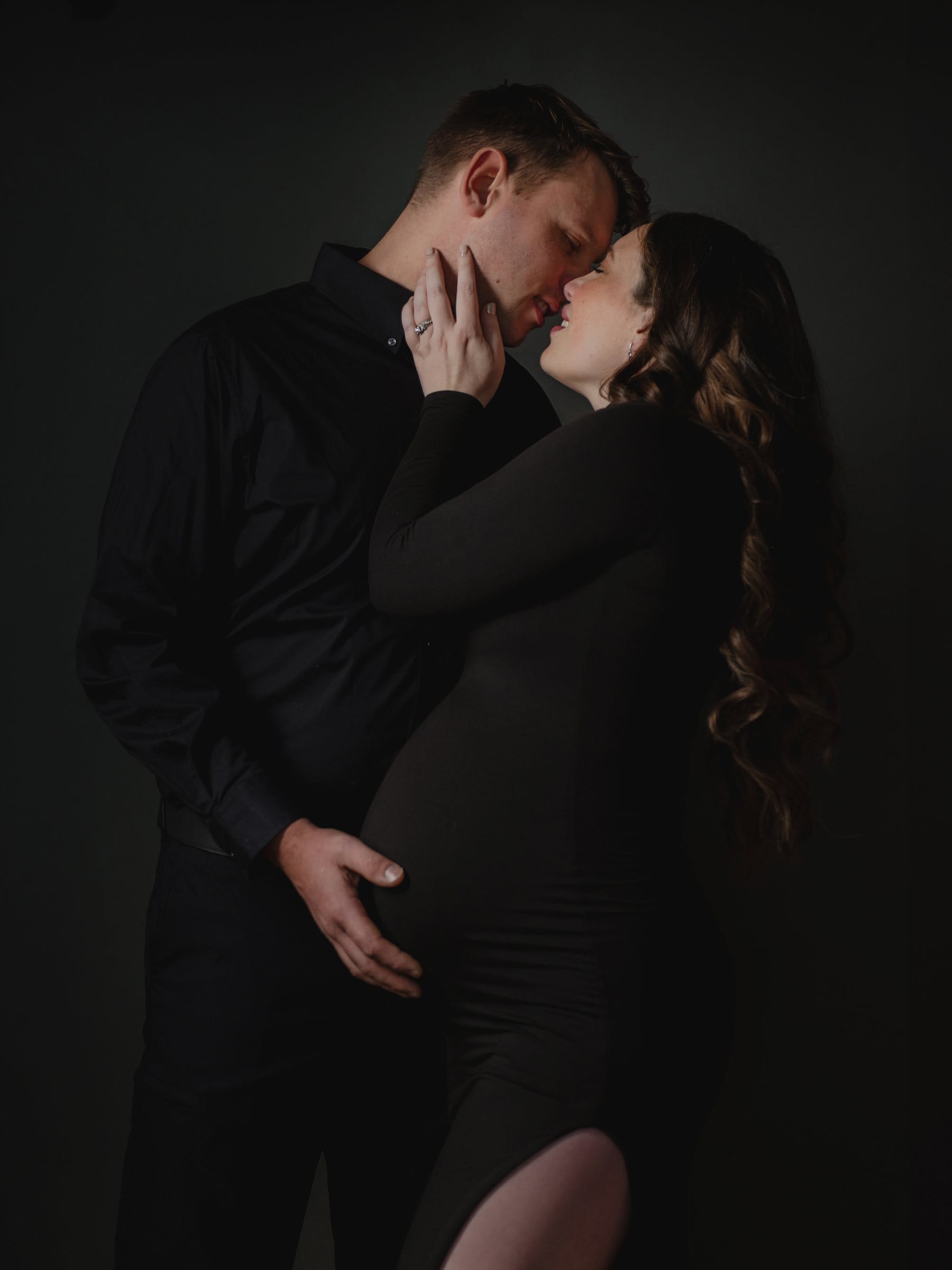 Expectant couple embracing against a black backdrop