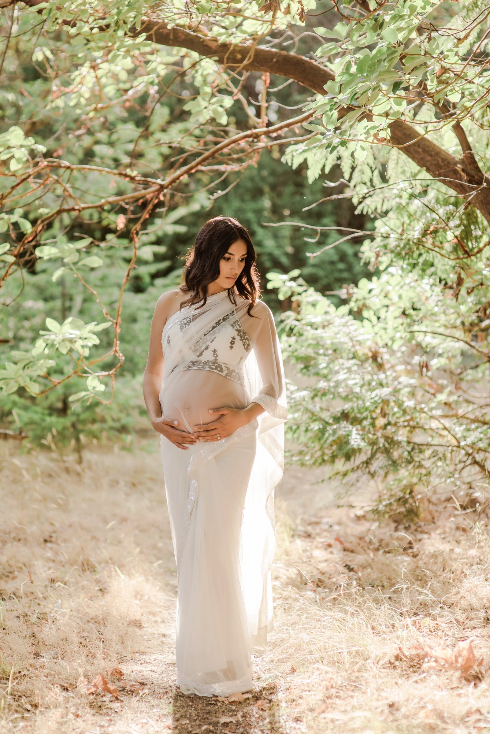 Expectant mother in a white gown in a sunlit woodland