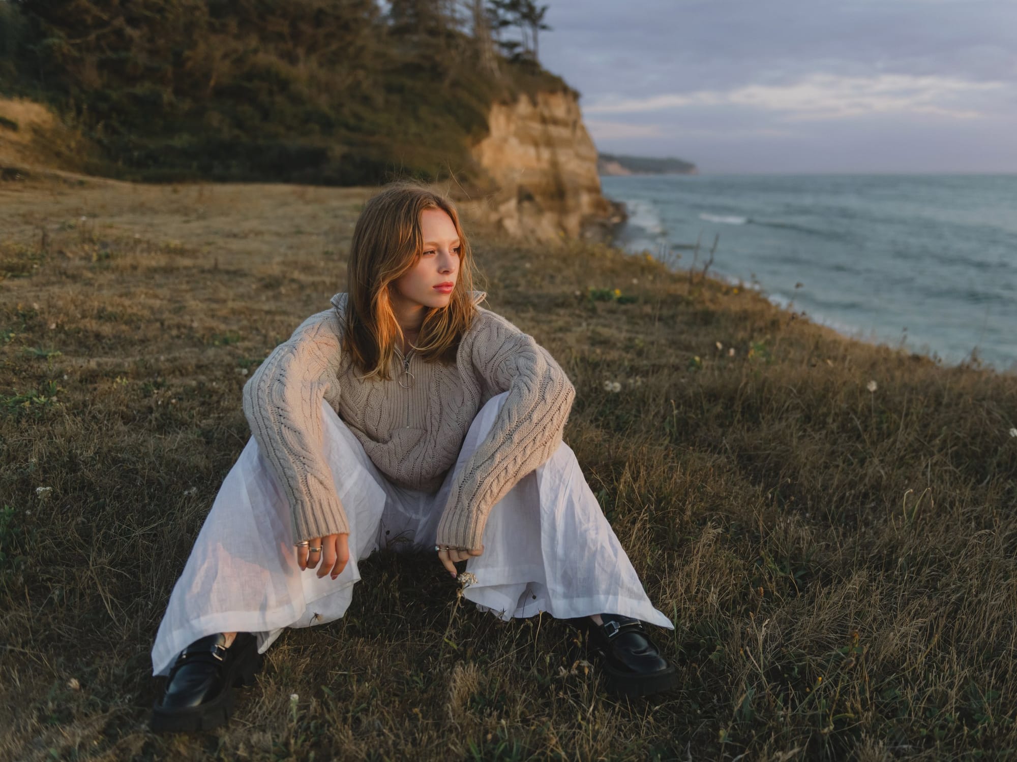 Young woman in a cream sweater sitting on a seaside bluff