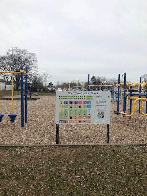 A blue and yellow playground is shown in the background. Forground is a large, printed AAC speech board also displaying a scannable QR code.