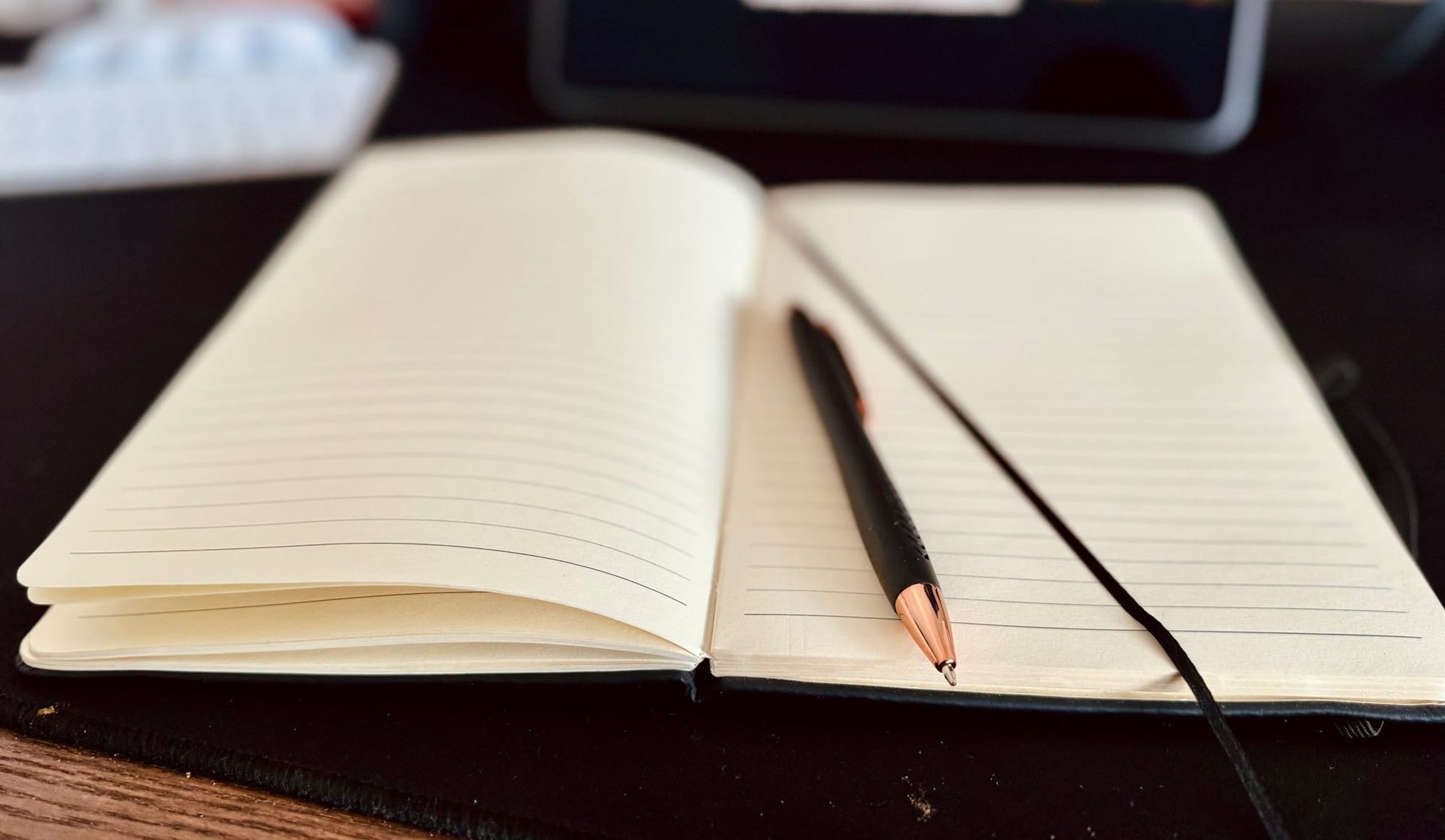 Open notebook with empty lined pages and a black pencil on a dark table, with a blurred tablet in the background.