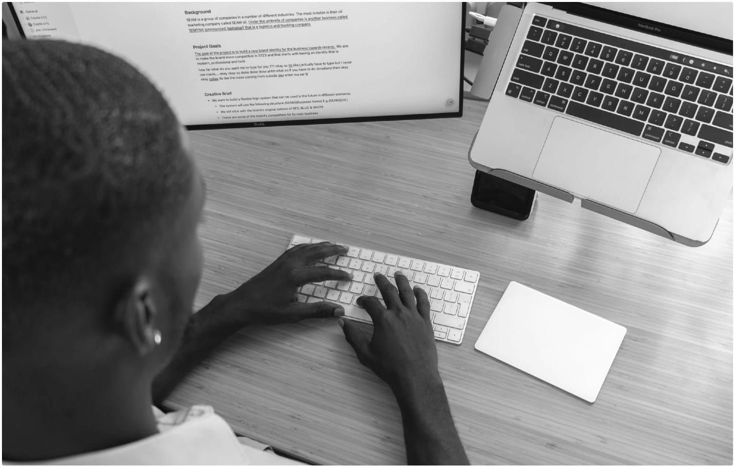 Person typing on a keyboard at a wooden desk, working with a large monitor and a laptop on a stand.