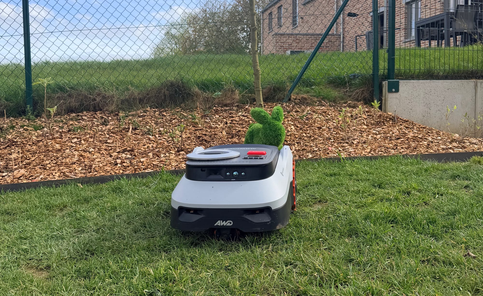 Navimow i210 LiDAR Pro en tonte le long d'un massif paillé avec un lapin décoratif en herbe synthétique posé derrière le robot