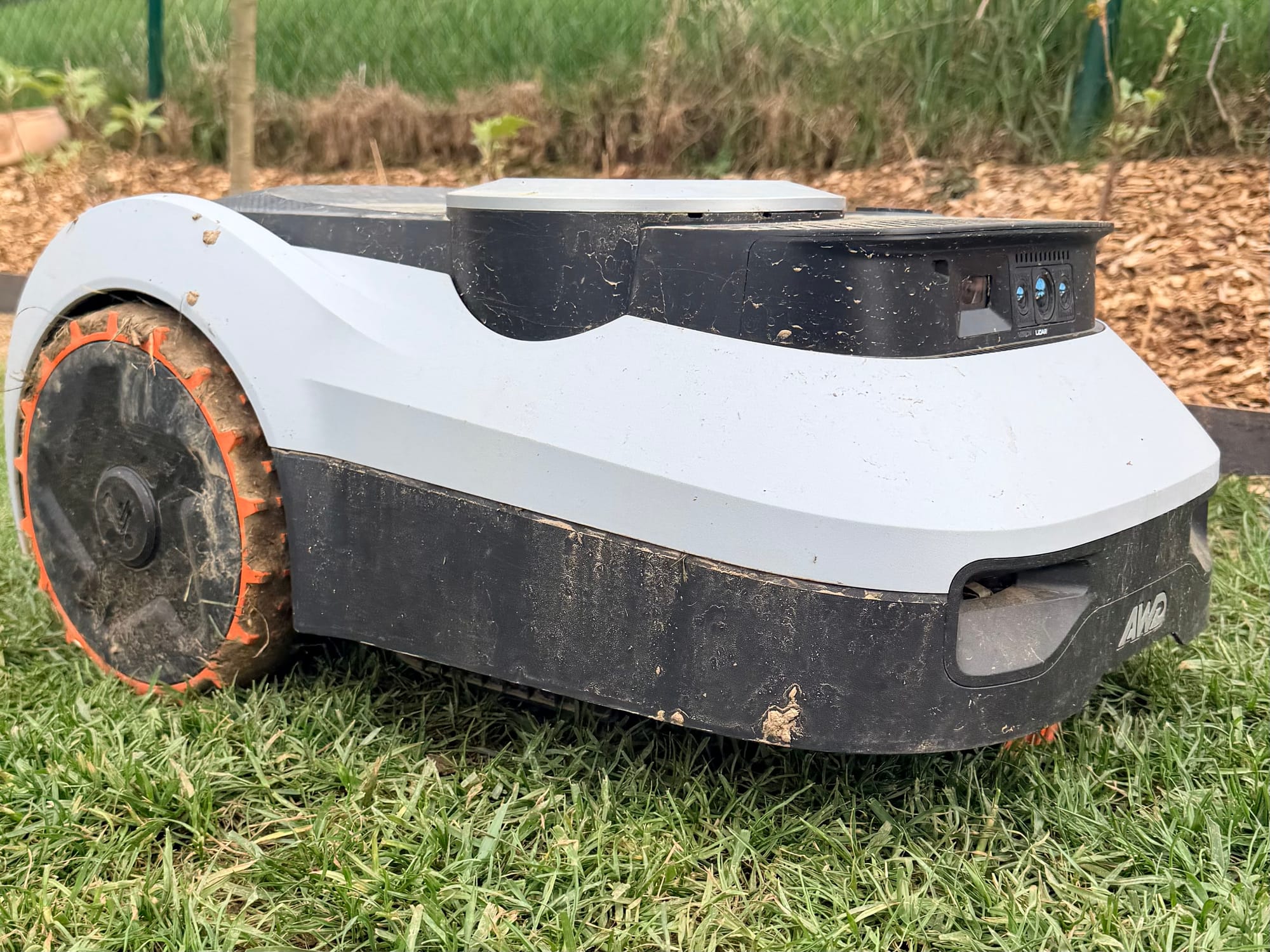 Navimow i210 LiDAR Pro vu de trois quarts après une session de tonte, carrosserie et roues encrassées d'herbe et de terre, capteurs Vision et LiDAR visibles