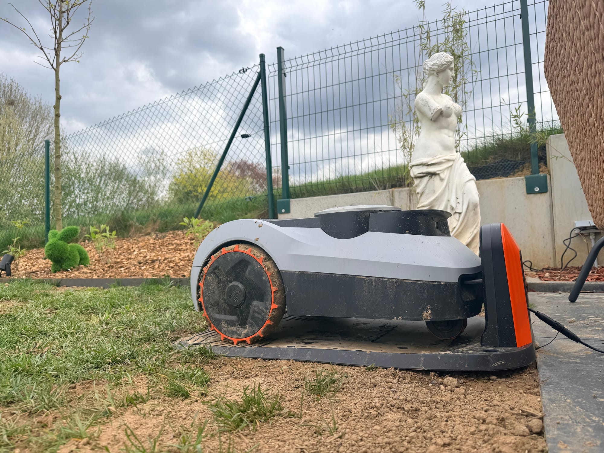 Navimow i210 LiDAR Pro sur sa station de recharge sans garage, roues encrassées de terre, sous un ciel nuageux dans le Brabant Wallon