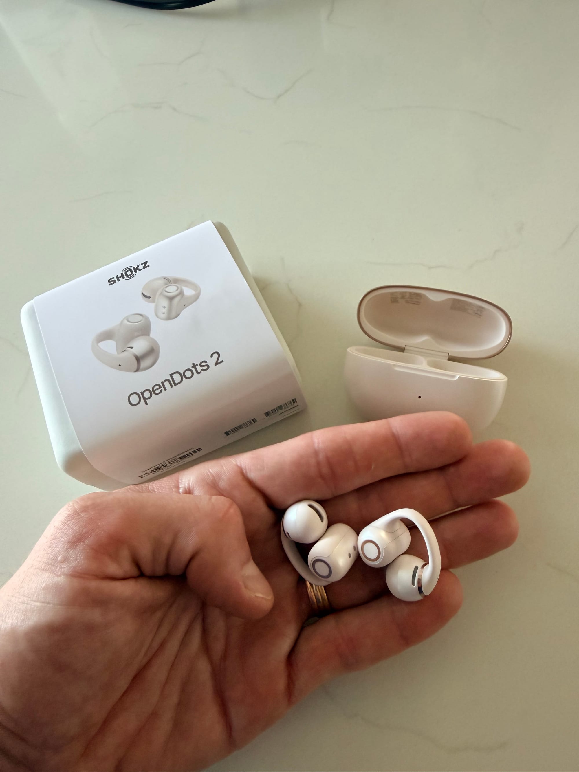 Shokz OpenDots 2 en pearl white dans la paume de la main, boîte et boîtier de charge en arrière-plan