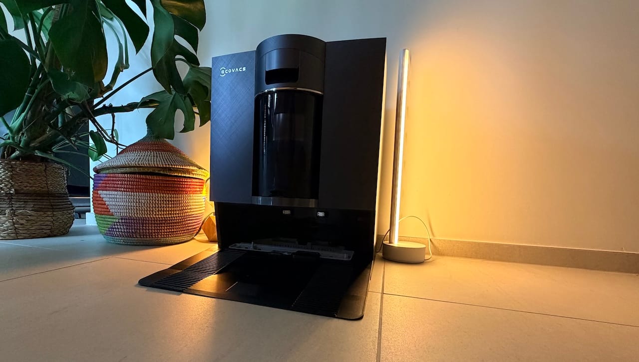 Station OmniCyclone de l'Ecovacs X12 dans un salon, finition noire avec réservoir visible, à côté d'un panier tressé et d'une lampe LED