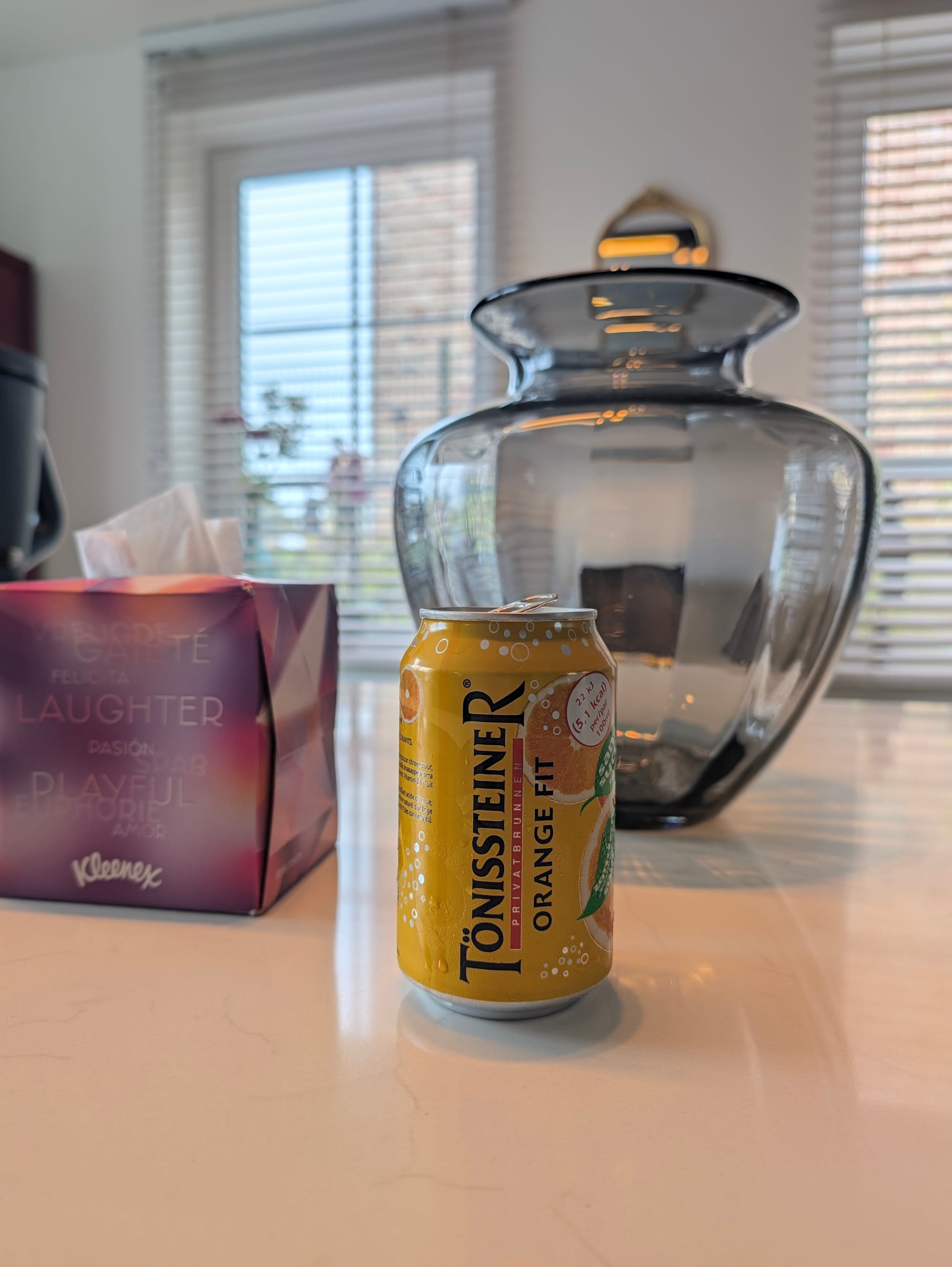 Photo prise avec le Pixel 11 Pro montrant une canette Tönissteiner Orange Fit avec bokeh naturel, vase en verre et stores en arrière-plan
