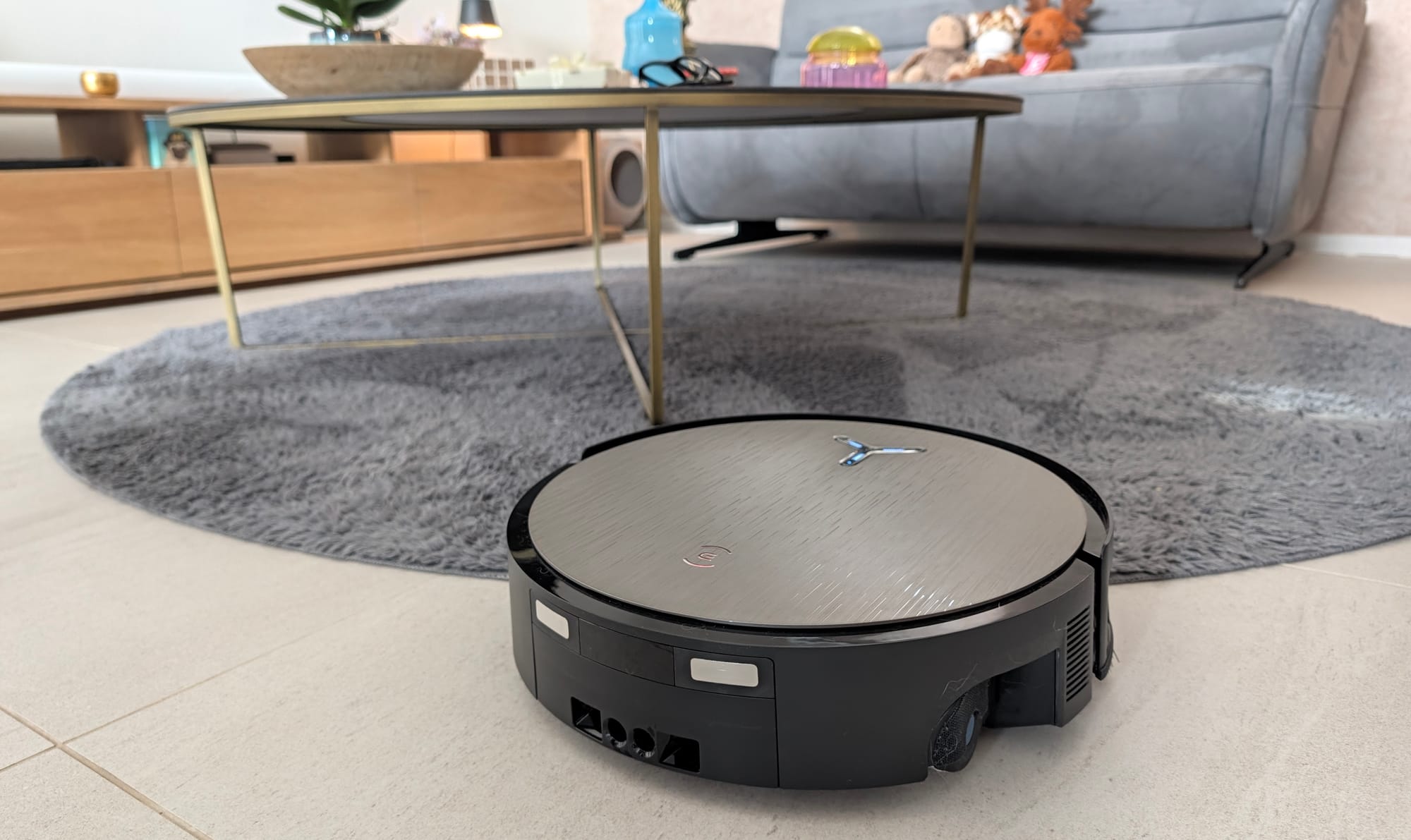 DEEBOT X12S OmniCyclone sur un tapis gris rond sous une table basse, jouets d'enfant et canapé en arrière-plan